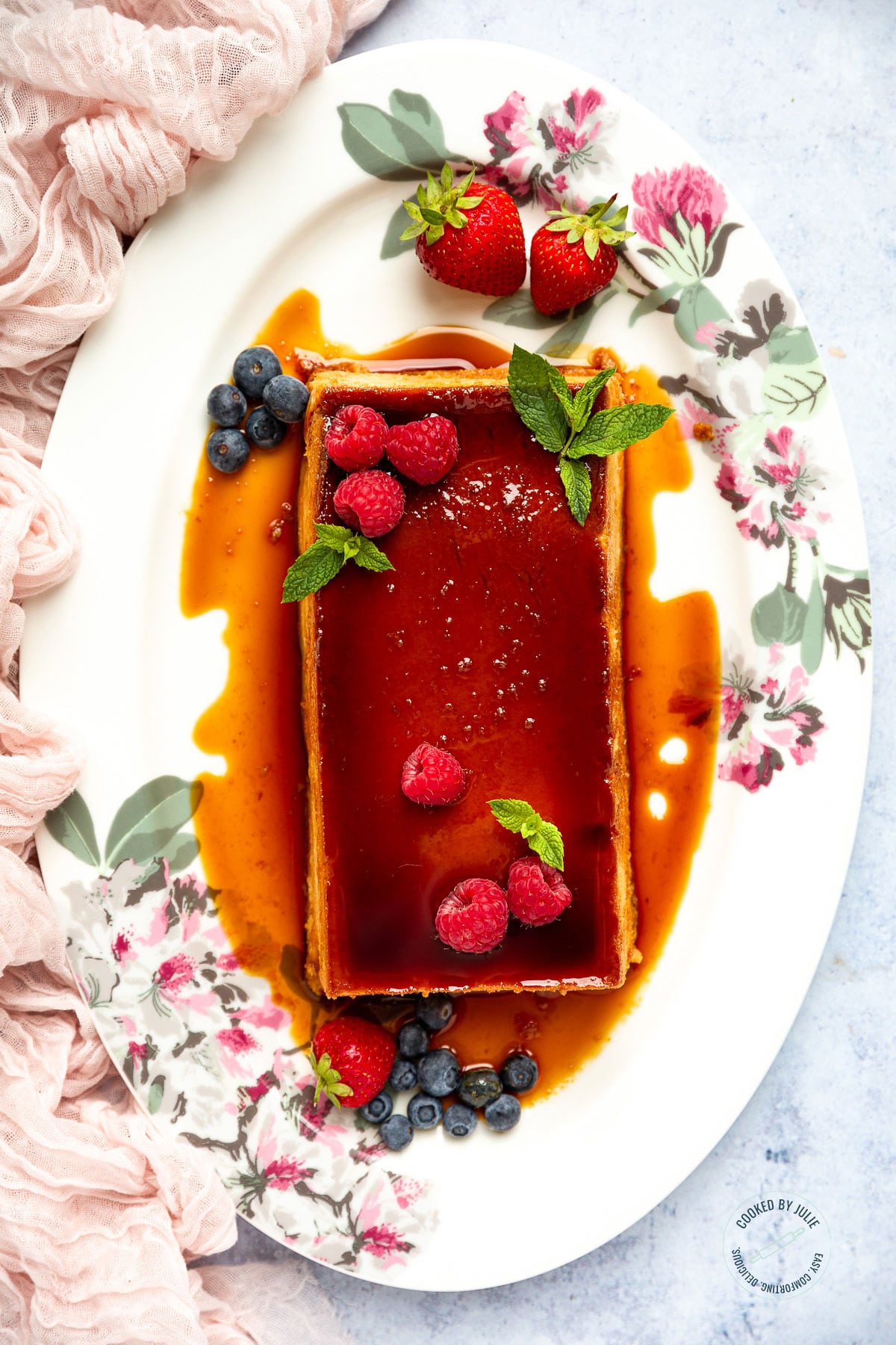flan with caramel, berries, and fresh mint on a white platter with a pink towel on the side. 