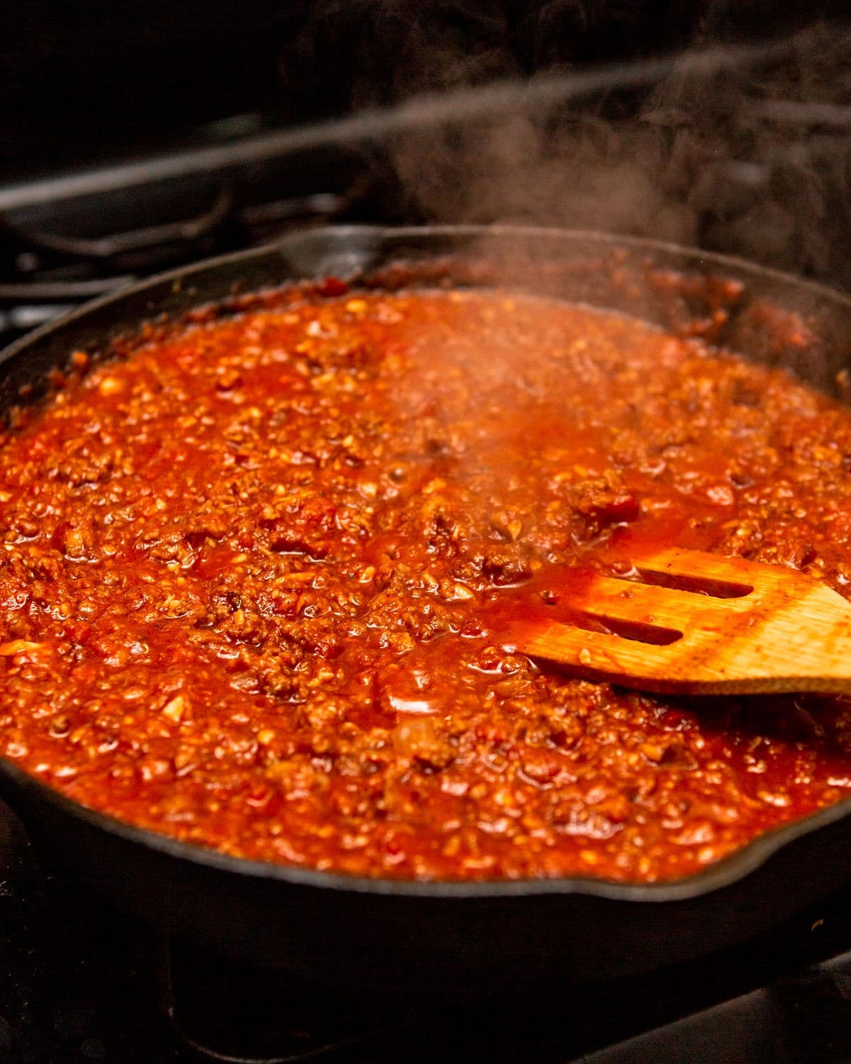 red meat sauce simmering in a cast iron skillet with a wooden spoon.