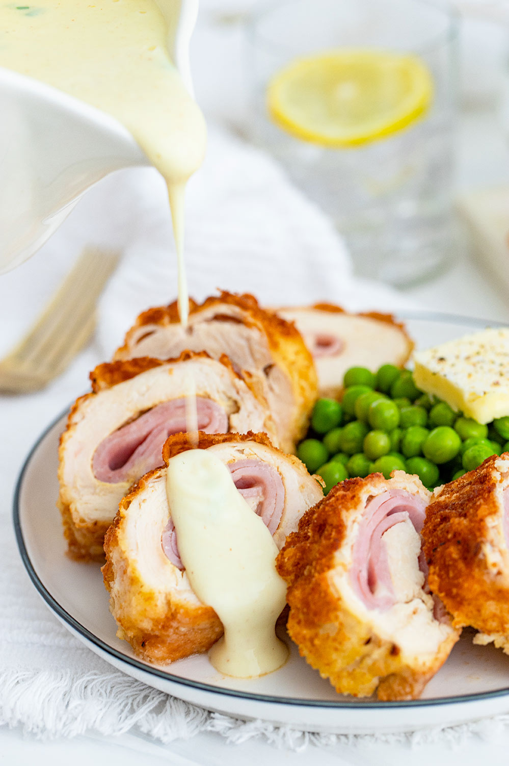chicken cordon bleu on a plate with sauce on top and green peas on the side.