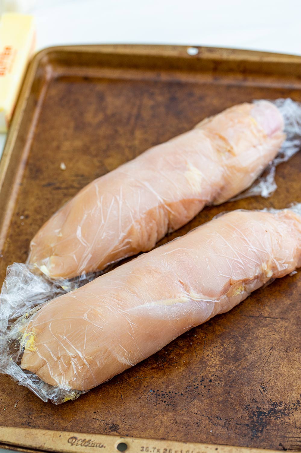 two chicken breasts rolled up in plastic on a baking sheet.