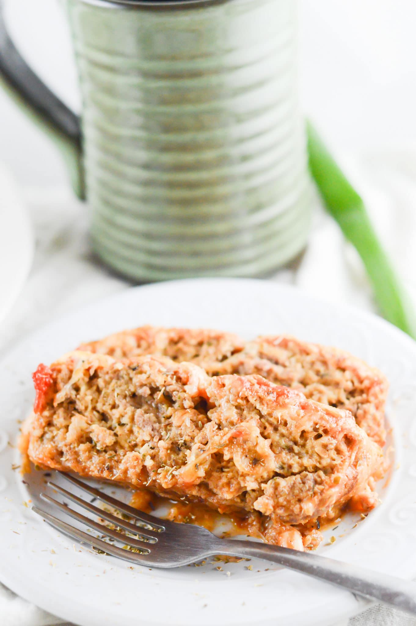 two slices of meatloaf on a white plate with a fork and a cup in the background. 