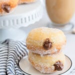 two nutella stuffed donuts on a small white plate with a blue and white towel on the side, a coffee and more donuts in the background.