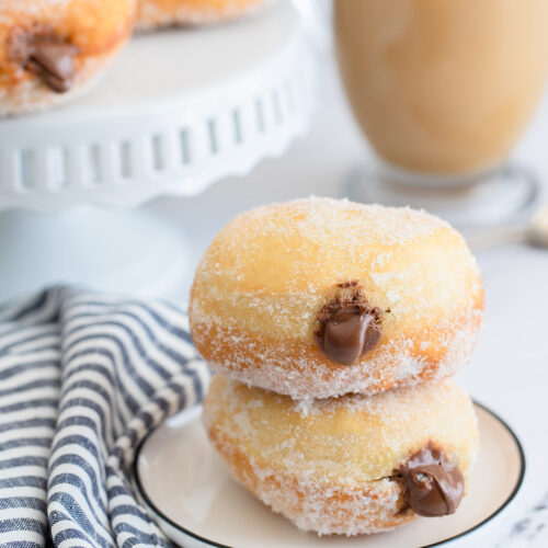 two nutella stuffed donuts on a small white plate with a blue and white towel on the side, a coffee and more donuts in the background.