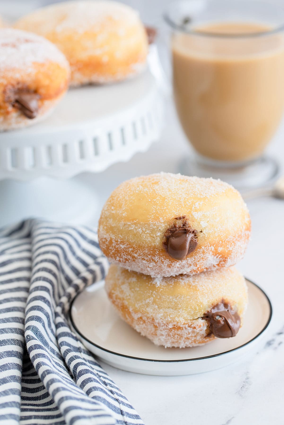 two nutella stuffed donuts on a small white plate with a blue and white towel on the side, a coffee and more donuts in the background.
