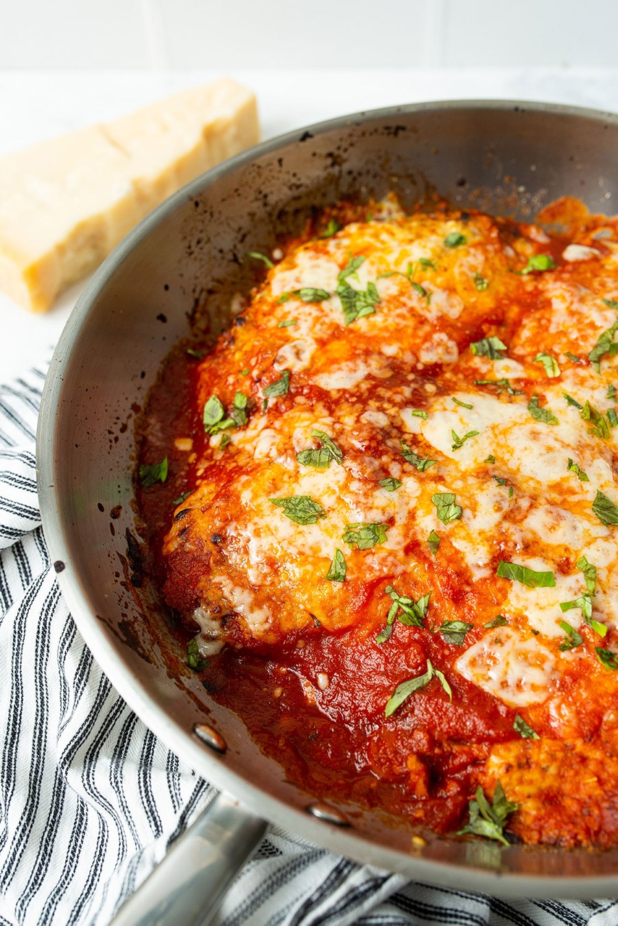 chicken parmesan in a skillet with tomato sauce and fresh mozzarella cheese. A blue and white towel on the side and a block of fresh parmesan cheese. 