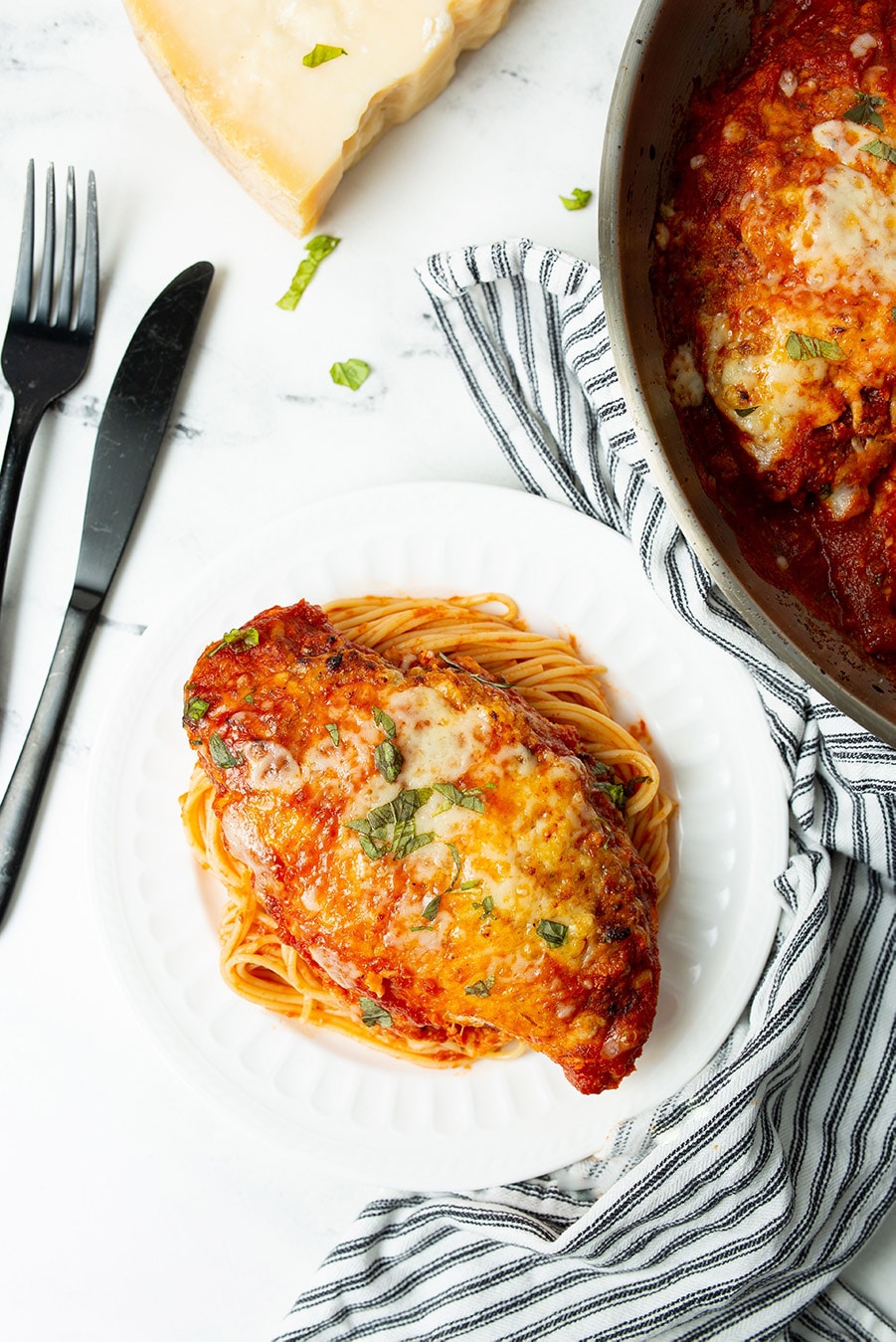 chicken parmigiana with spaghetti on a white plate with a knife and fork on the side and a blue and white towel. 