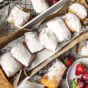 a long bog with brown paper and beignets inside. beignets on the side of the box along with strawberries and blueberries.