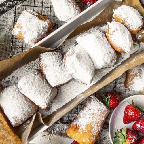 a long bog with brown paper and beignets inside. beignets on the side of the box along with strawberries and blueberries.