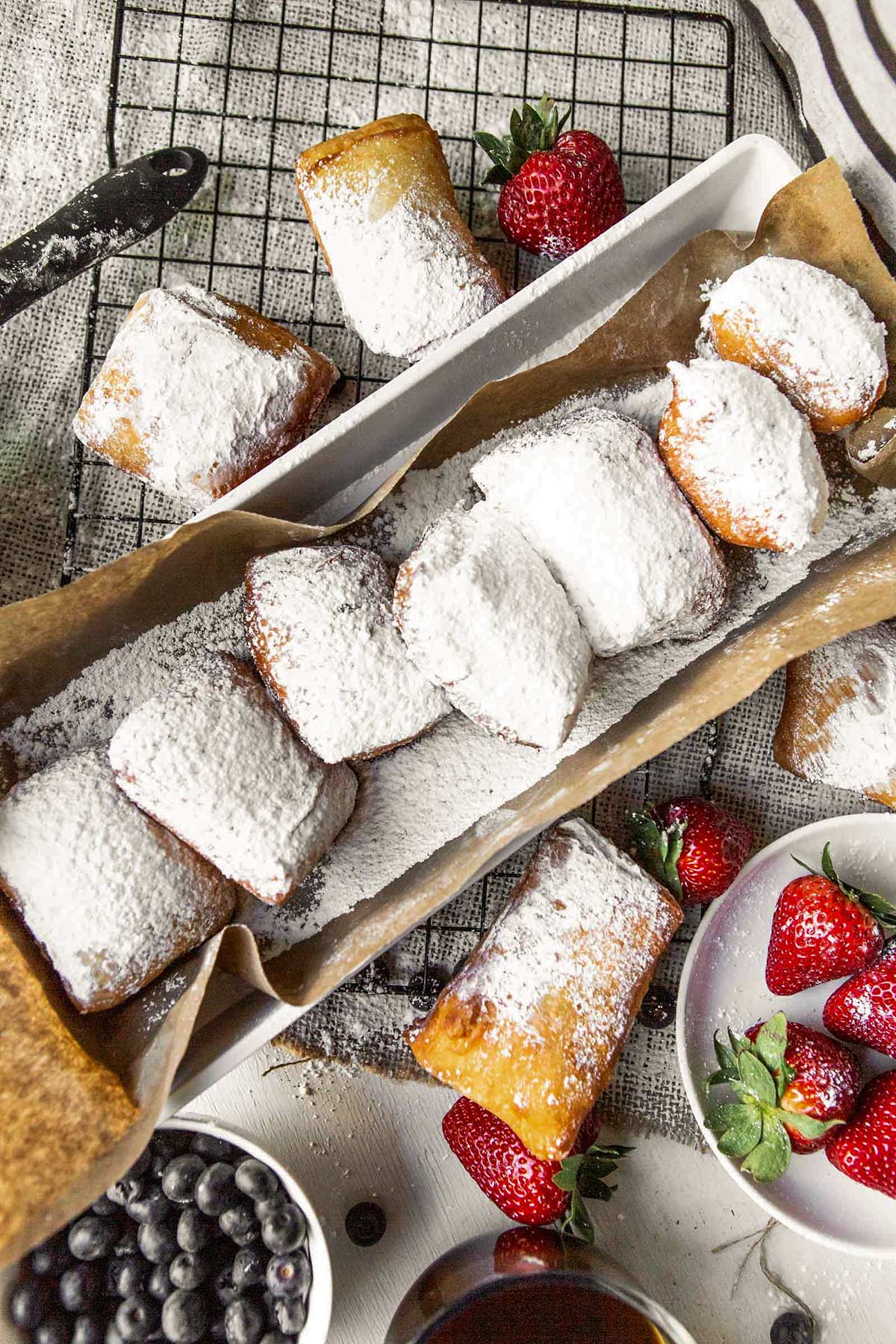 a long bog with brown paper and beignets inside. beignets on the side of the box along with strawberries and blueberries.