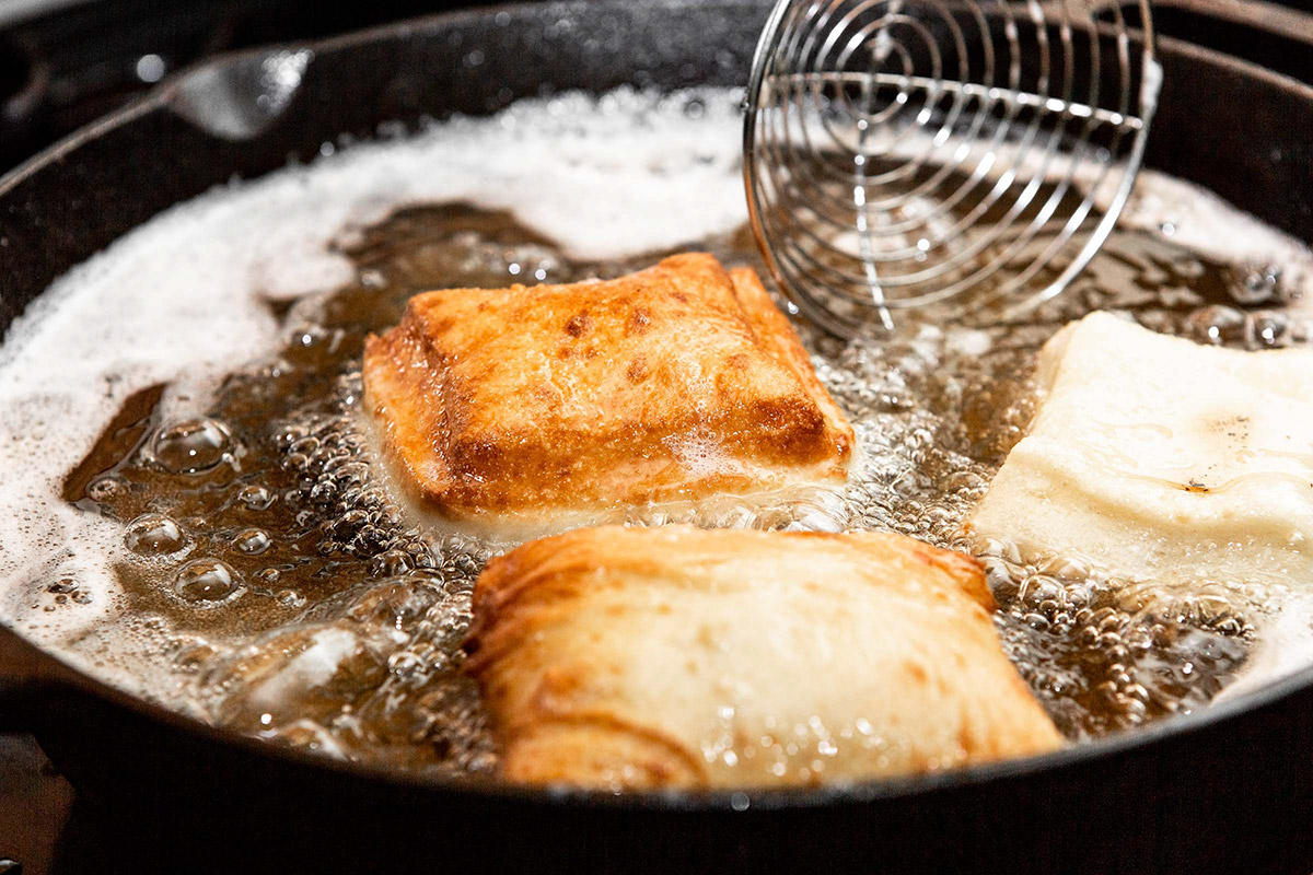 three beignets frying in a skillet with oil and a round mesh skimmer.