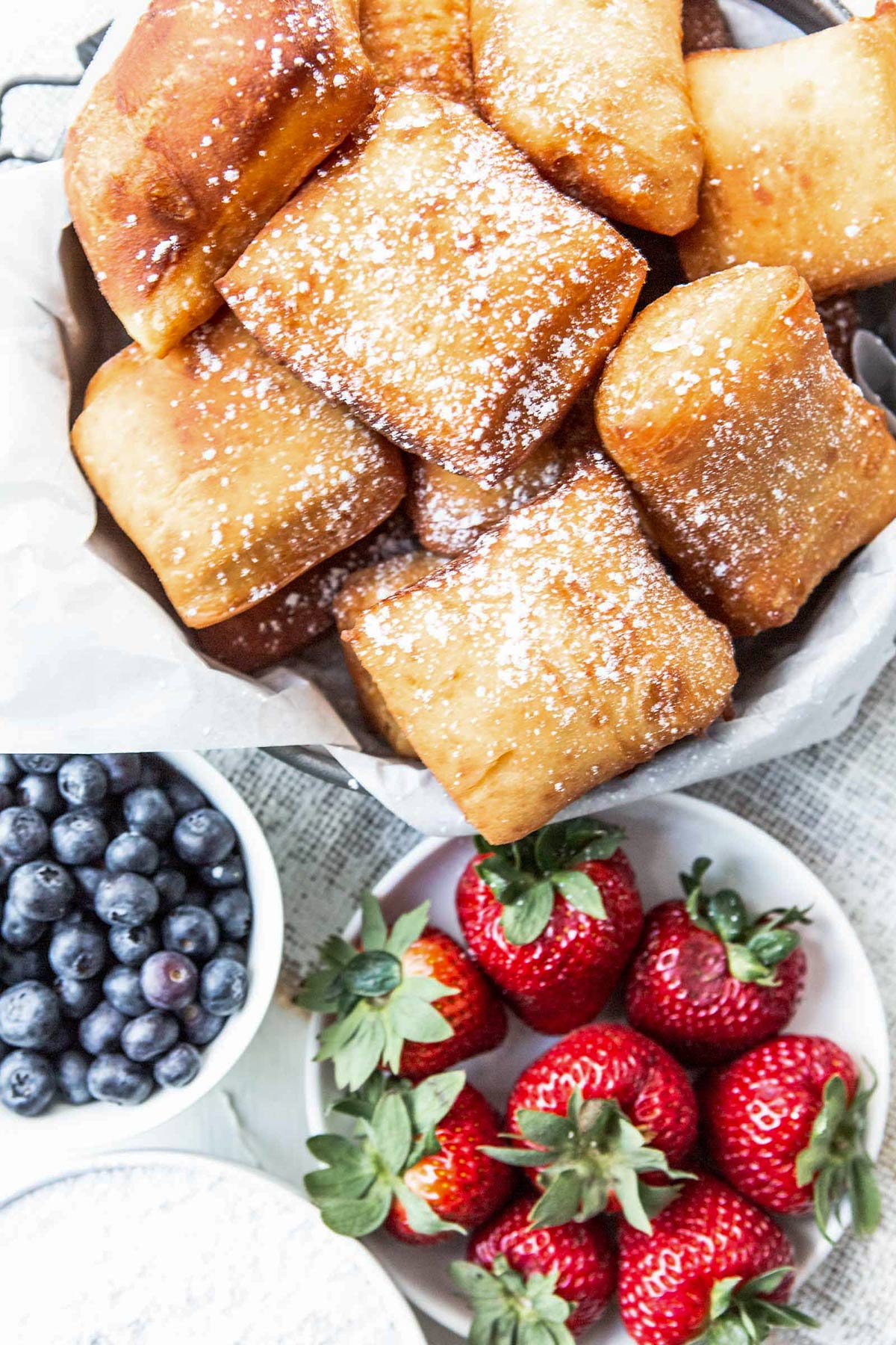 beignets with powdered sugar on top inside of a bowl with two small bowls on the side filled with strawberries and blueberries.