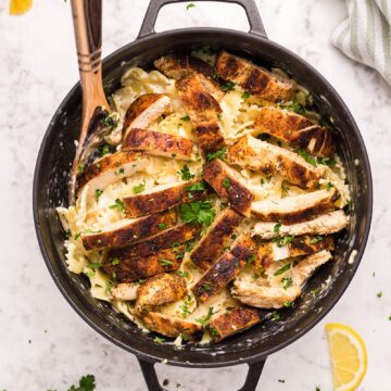 Cajun chicken, farfalle pasta, parsley, and a wooden spoon in a black skillet.