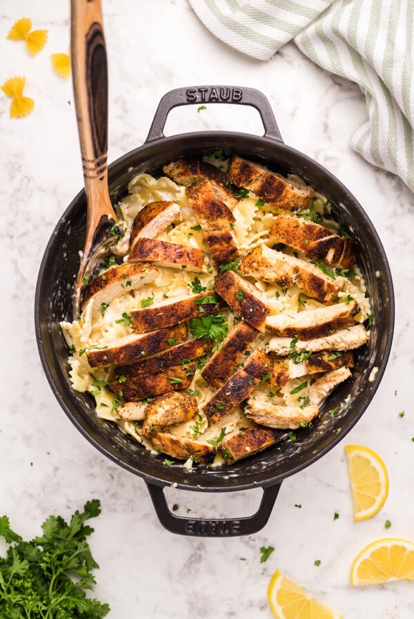 Cajun chicken, farfalle pasta, parsley, and a wooden spoon in a black skillet.