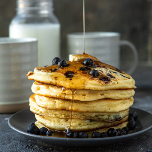 six blueberry pancakes on a blue plate with butter and maple syrup on top. A jug of milk in the background with two mugs and fresh blueberries.