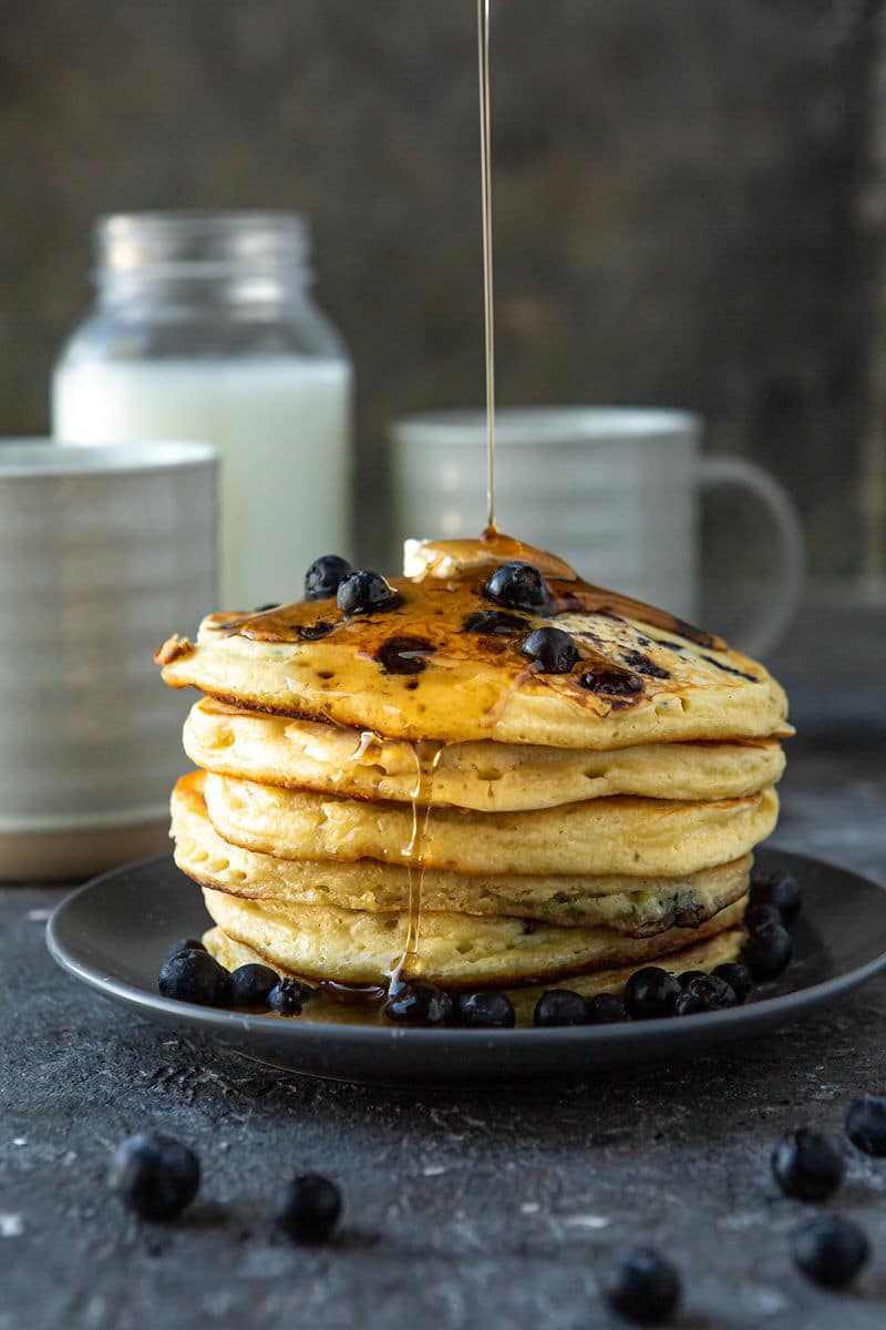 six blueberry pancakes on a blue plate with butter and maple syrup on top. A jug of milk in the background with two mugs and fresh blueberries.