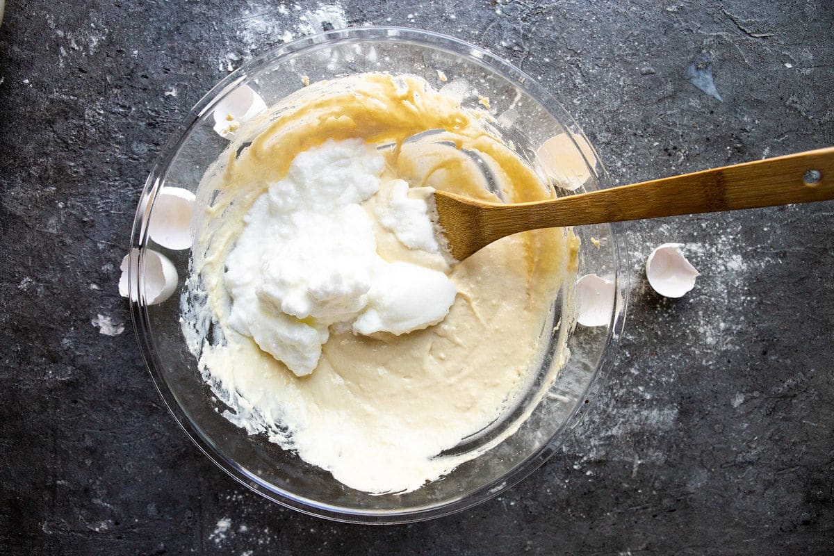 A bowl with pancake batter and folded egg whites with a spatula. Egg shells on the side of the bowl. 