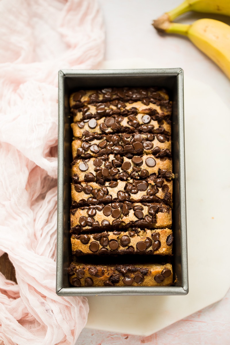 banana bread in a loaf pan cut into slices with bananas and a pink towel on the side