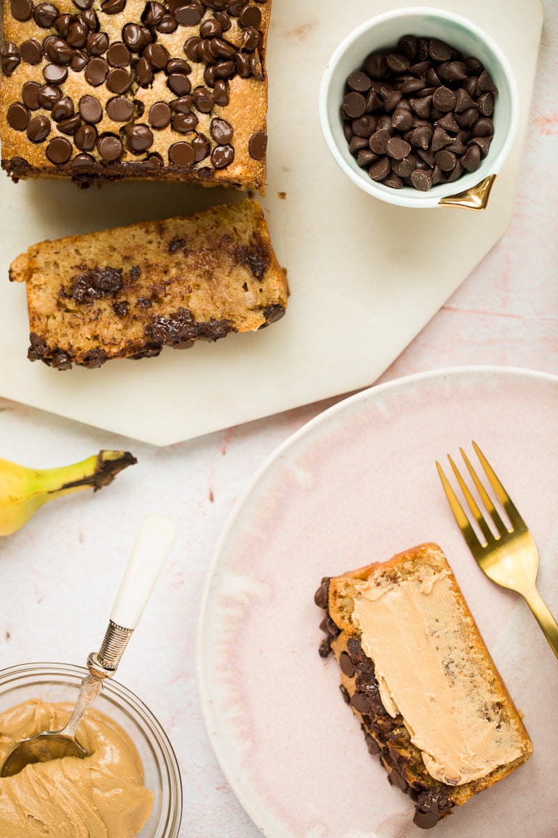 banana bread, chocolate chips in a small bowl, a banana, peanut butter in a bowl, and a slice of banana bread with smeared peanut butter and a fork on the side.