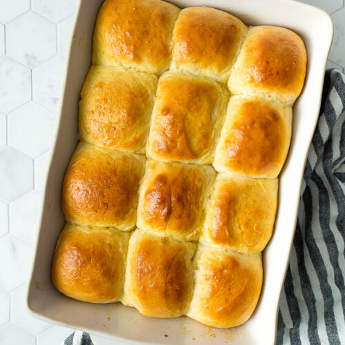 a dozen baked dinner rolls in a white baking dish on top of a white and blue kitchen towel.