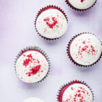 Six red velvet cupcakes with cream cheese frosting and cupcake crumbs on top.