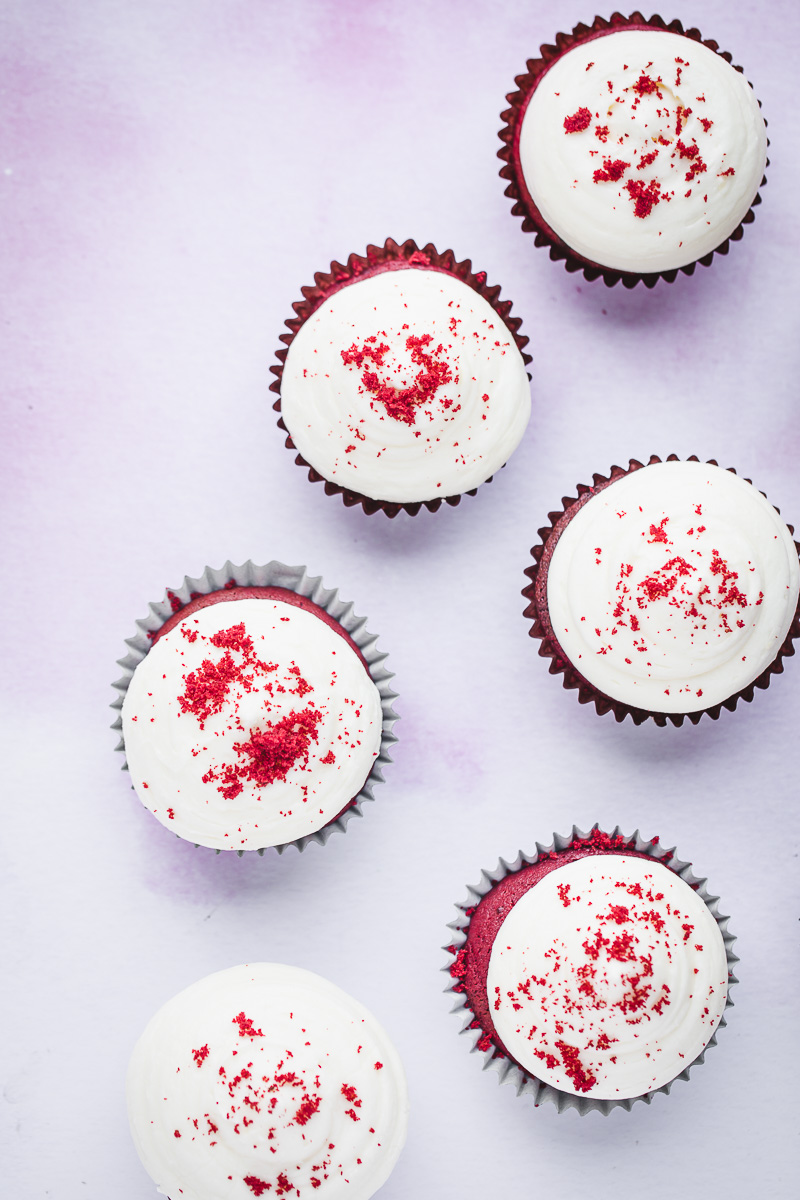 Six red velvet cupcakes with cream cheese frosting and cupcake crumbs on top. 