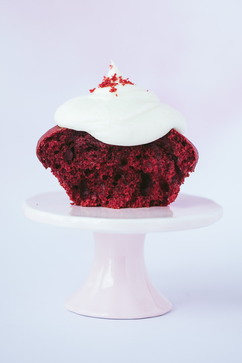 one red velvet cupcake with cream cheese frosting sliced in half on top of a pink cake stand. 