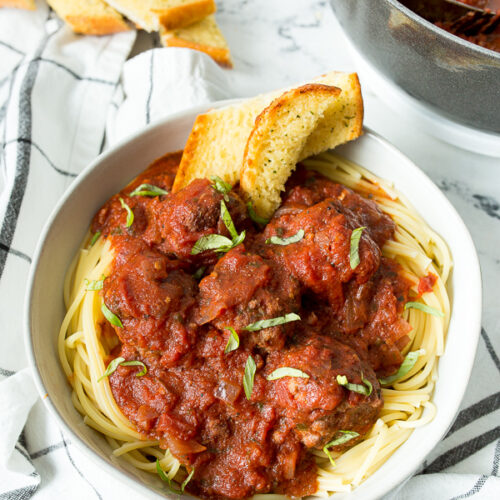 a white bowl filled with spaghetti and meatballs, garlic bread on the side, a white and blue towel underneath the bowl, and a pot of meatballs in the background.