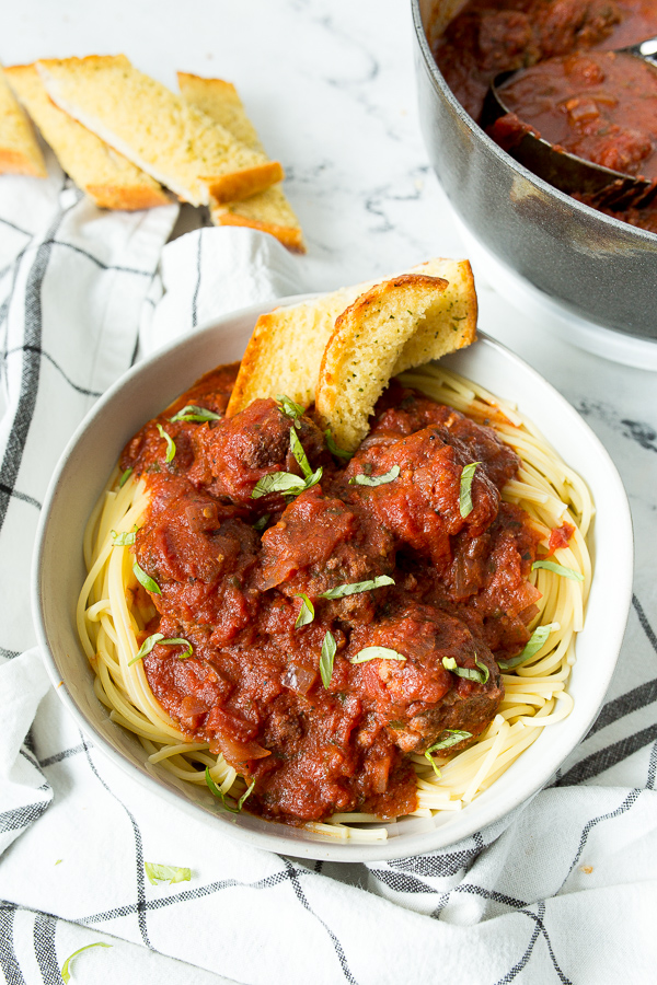 a white bowl filled with spaghetti and meatballs, garlic bread on the side, a white and blue towel underneath the bowl, and a pot of meatballs in the background. 