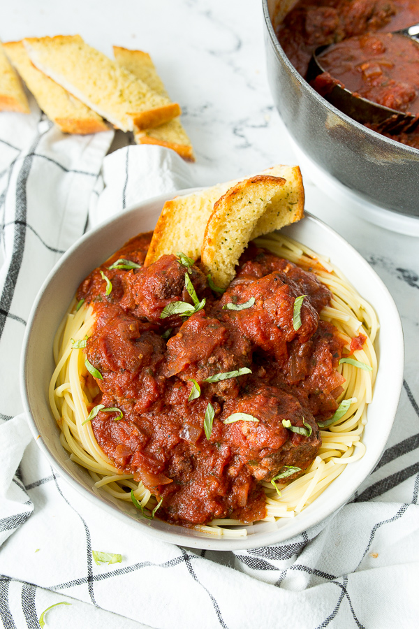 a white bowl filled with spaghetti and meatballs, garlic bread on the side, a white and blue towel underneath the bowl, and a pot of meatballs in the background.