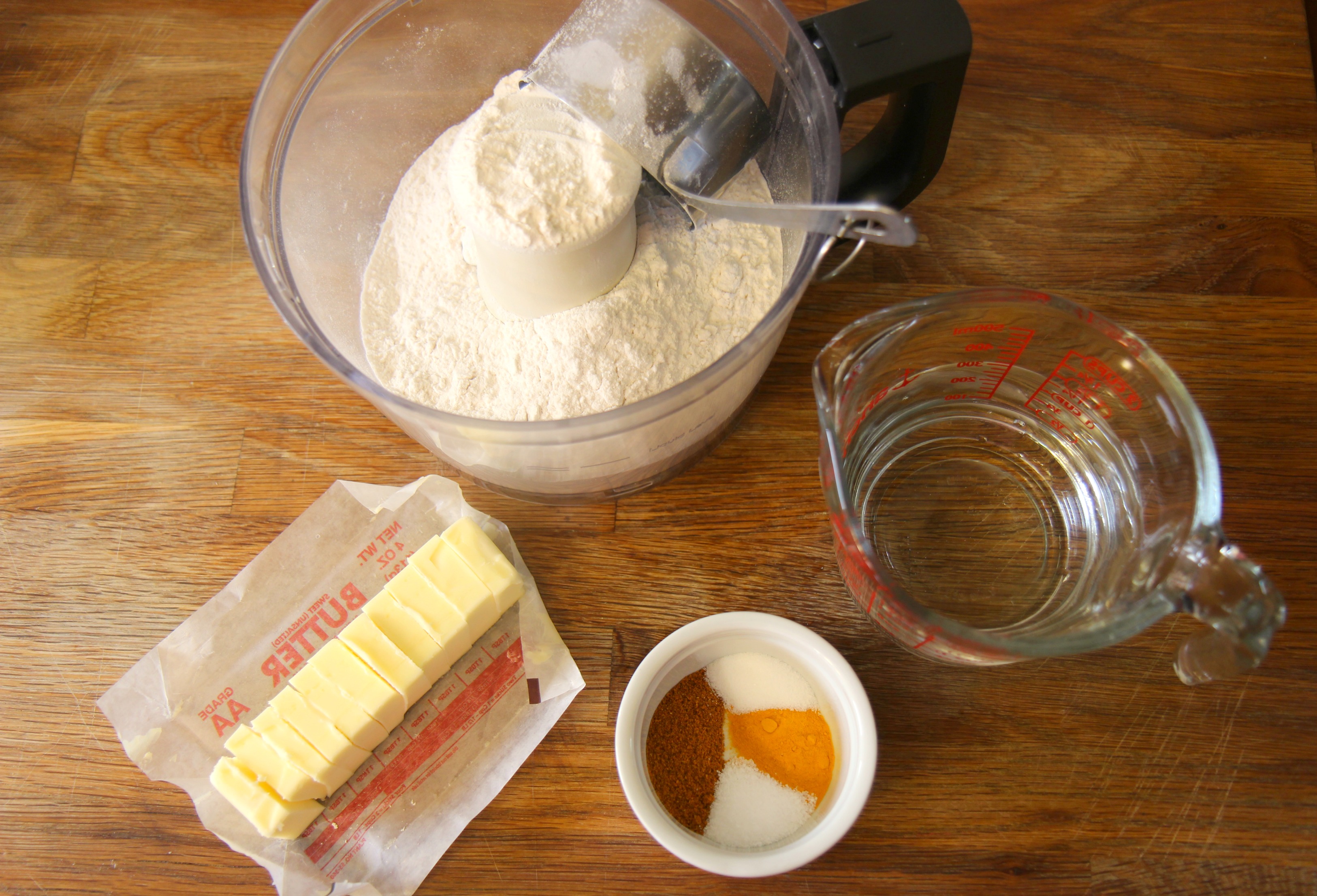 flour in a food processor, stick of butter, spices in a small ramekin, and a measuring cup with water. 
