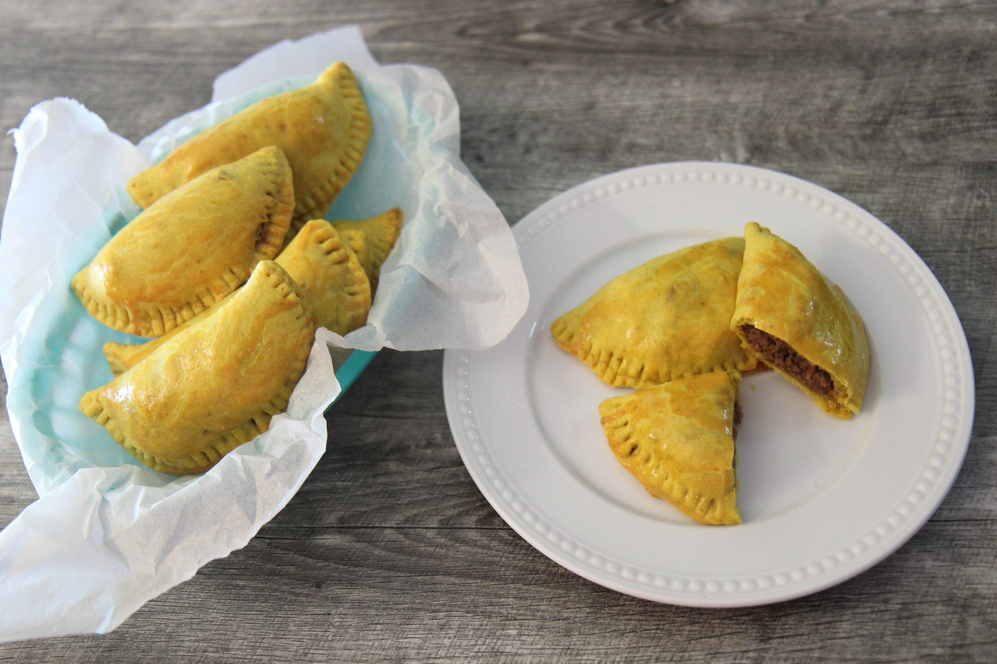 five Jamaican beef patties in a blue basket and two on a white plate. 