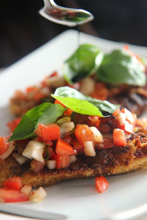 fried chicken breasts with fresh basil, tomatoes, and onions on a white plate. A spoon drizzling balsamic over the chicken.
