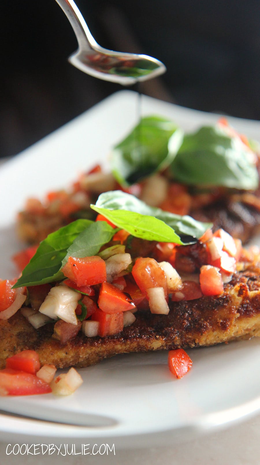 fried chicken breasts with fresh basil, tomatoes, and onions on a white plate. A spoon drizzling balsamic over the chicken.