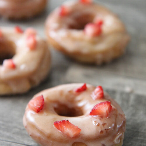 four fresh strawberry donuts on a gray surface.