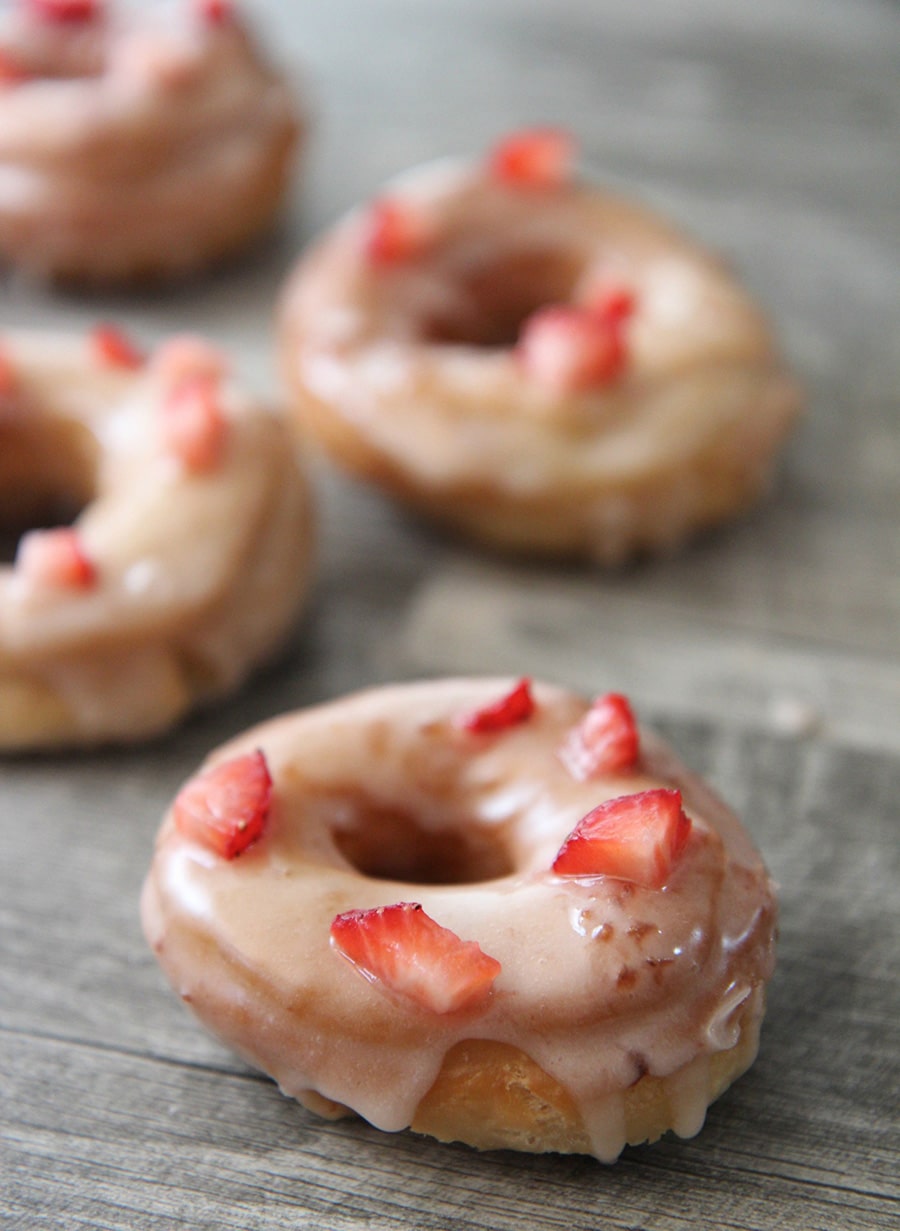 four fresh strawberry donuts on a gray surface.