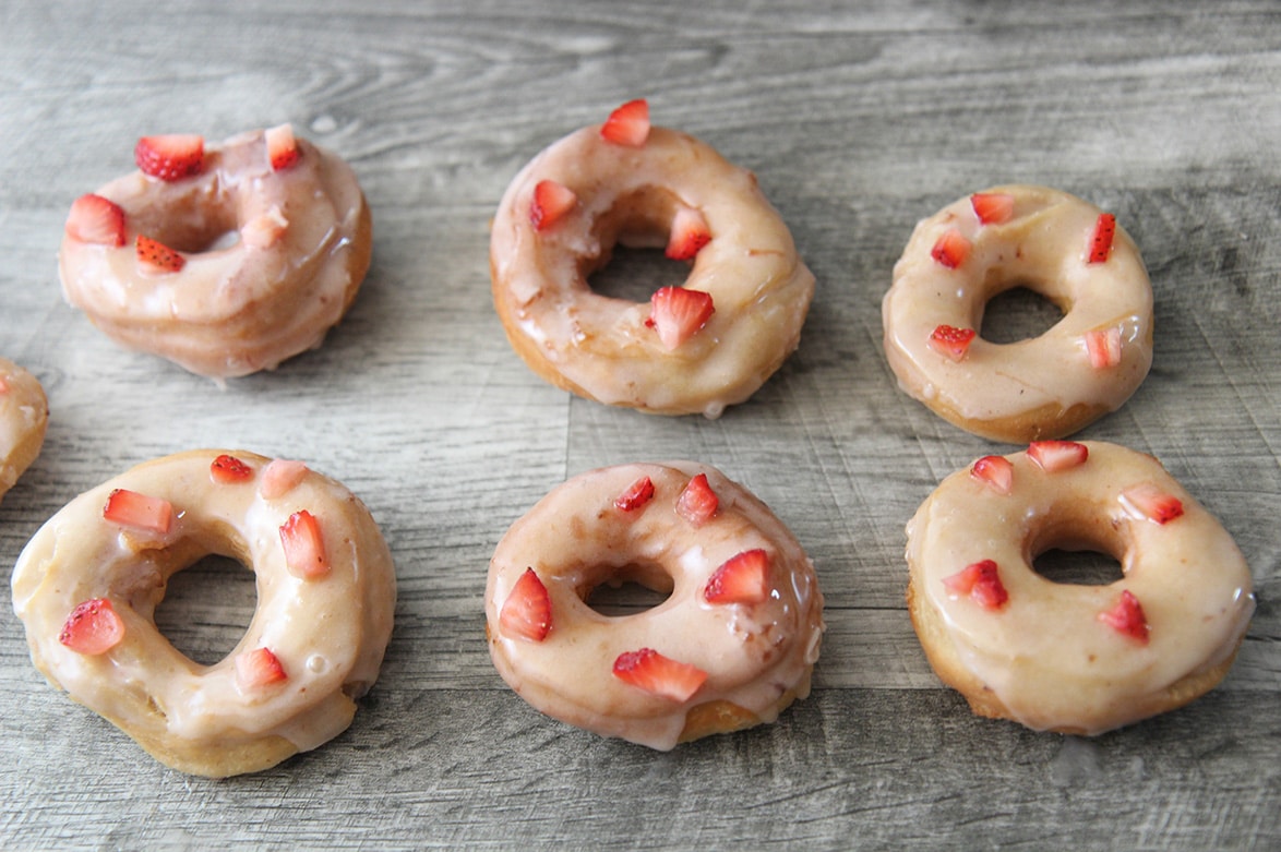 six glazed strawberry donuts on a gray surface.