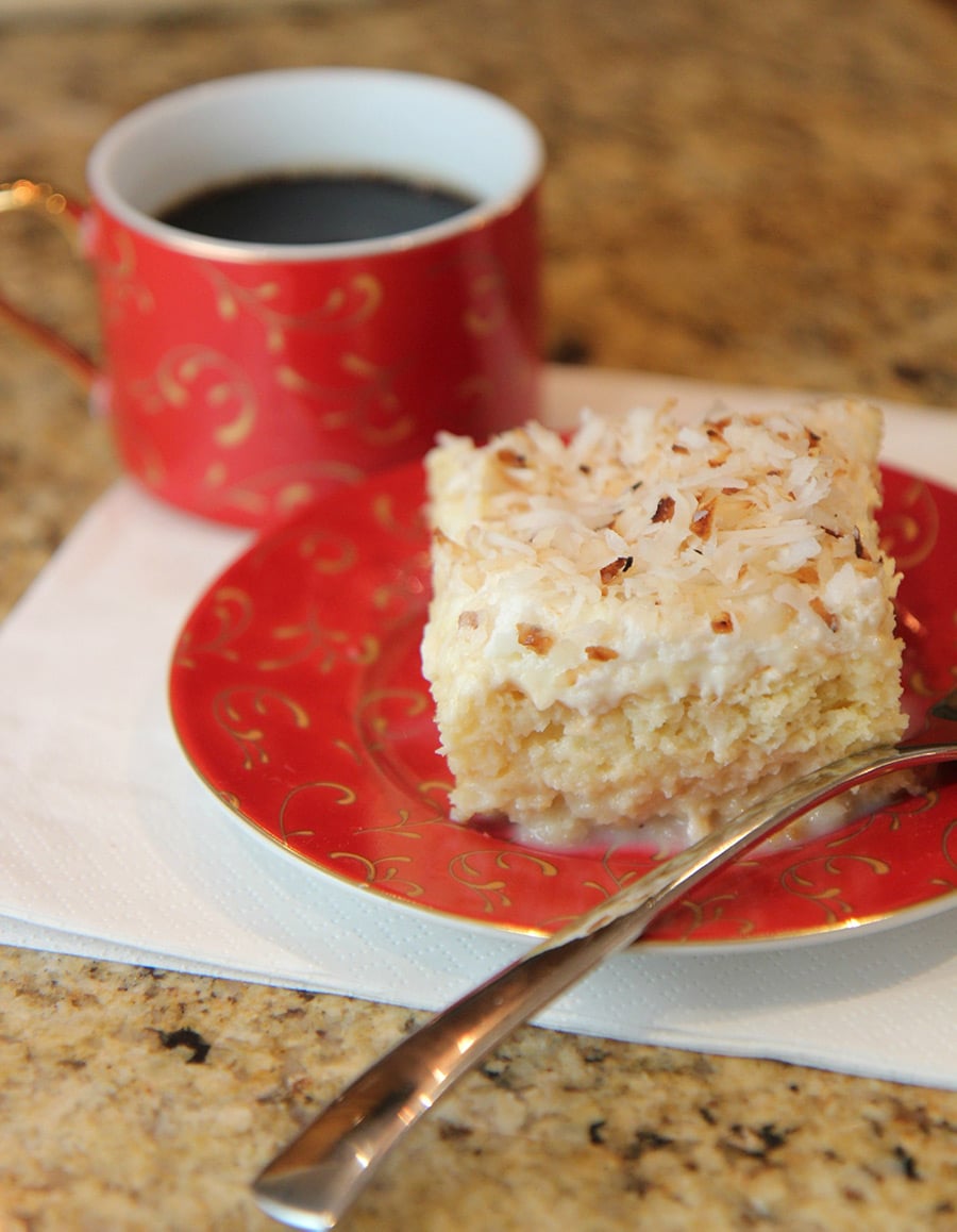 a slice of tres leches coconut cake on a red small plate with a cup of coffee on the side and a fork.