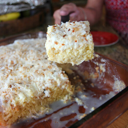 a hand lifting a slice of coconut tres leches and a full cake in the background.