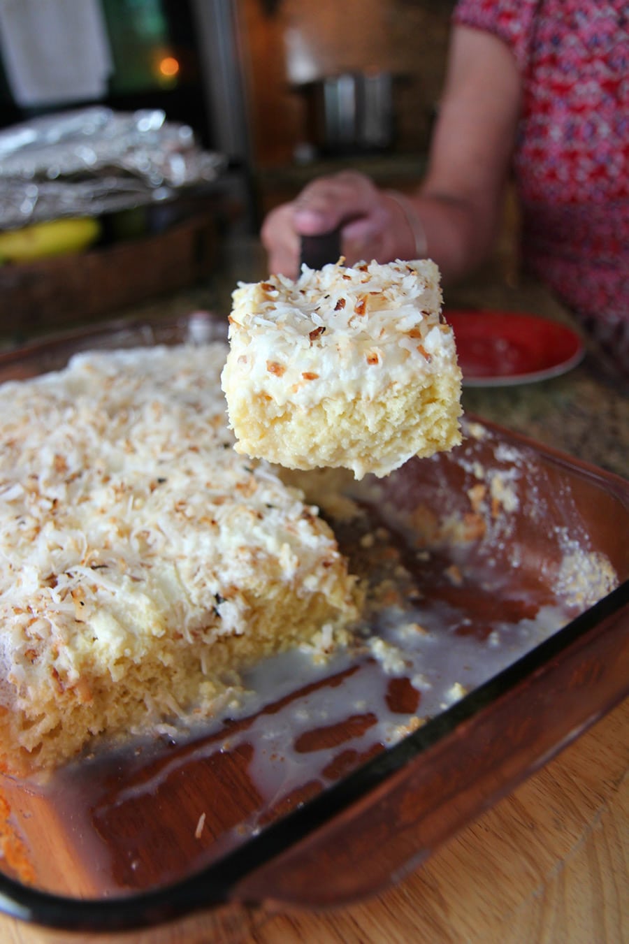 a hand lifting a slice of coconut tres leches and a full cake in the background.