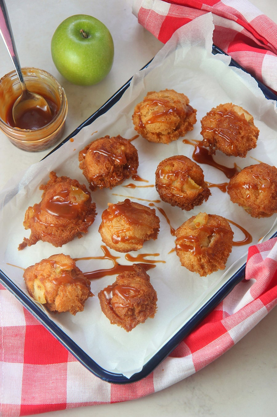 ten fried apple fritters with caramel sauce on top. A small jar of caramel on the side, a green apple, and a red and white towel.. 