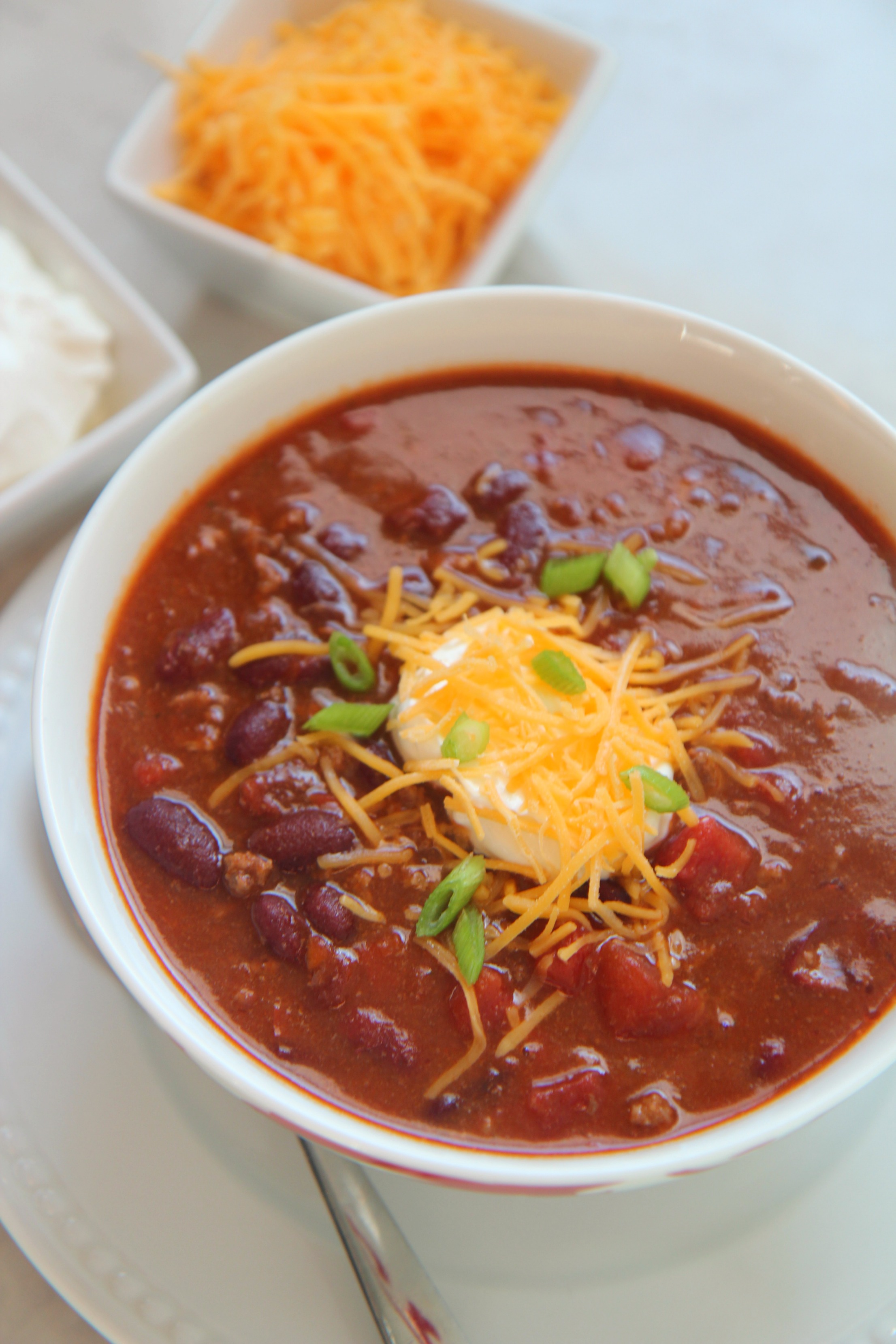 Chili with sour cream, cheddar cheese, and scallions in a white bowl with a spoon, cheese, and sour cream on the side. 