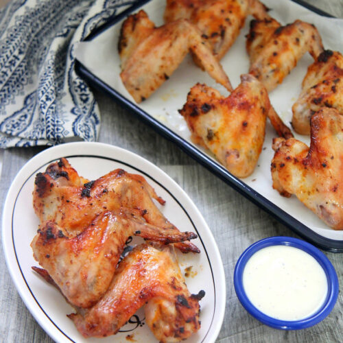 oven roasted chicken wings on a small white plate with a side of blue cheese dip and more chicken wings in the background.