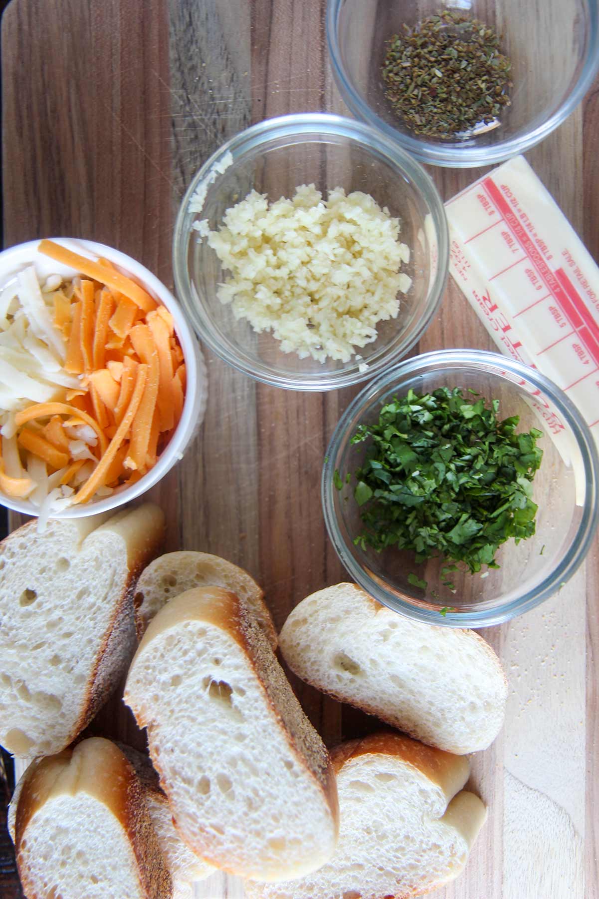 cheese, garlic, and herbs in small bowls and bread and butter on a wooden board. 