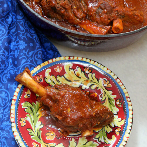 braised lamb shank of a red and yellow plate with a red pot in the background with lamb and a blue towel on the side.