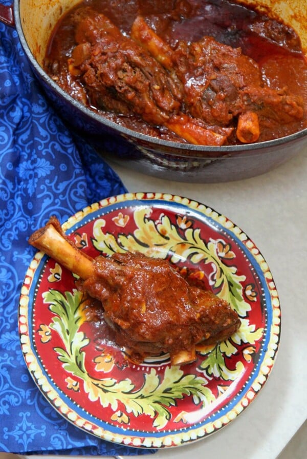 braised lamb shank of a red and yellow plate with a red pot in the background with lamb and a blue towel on the side.