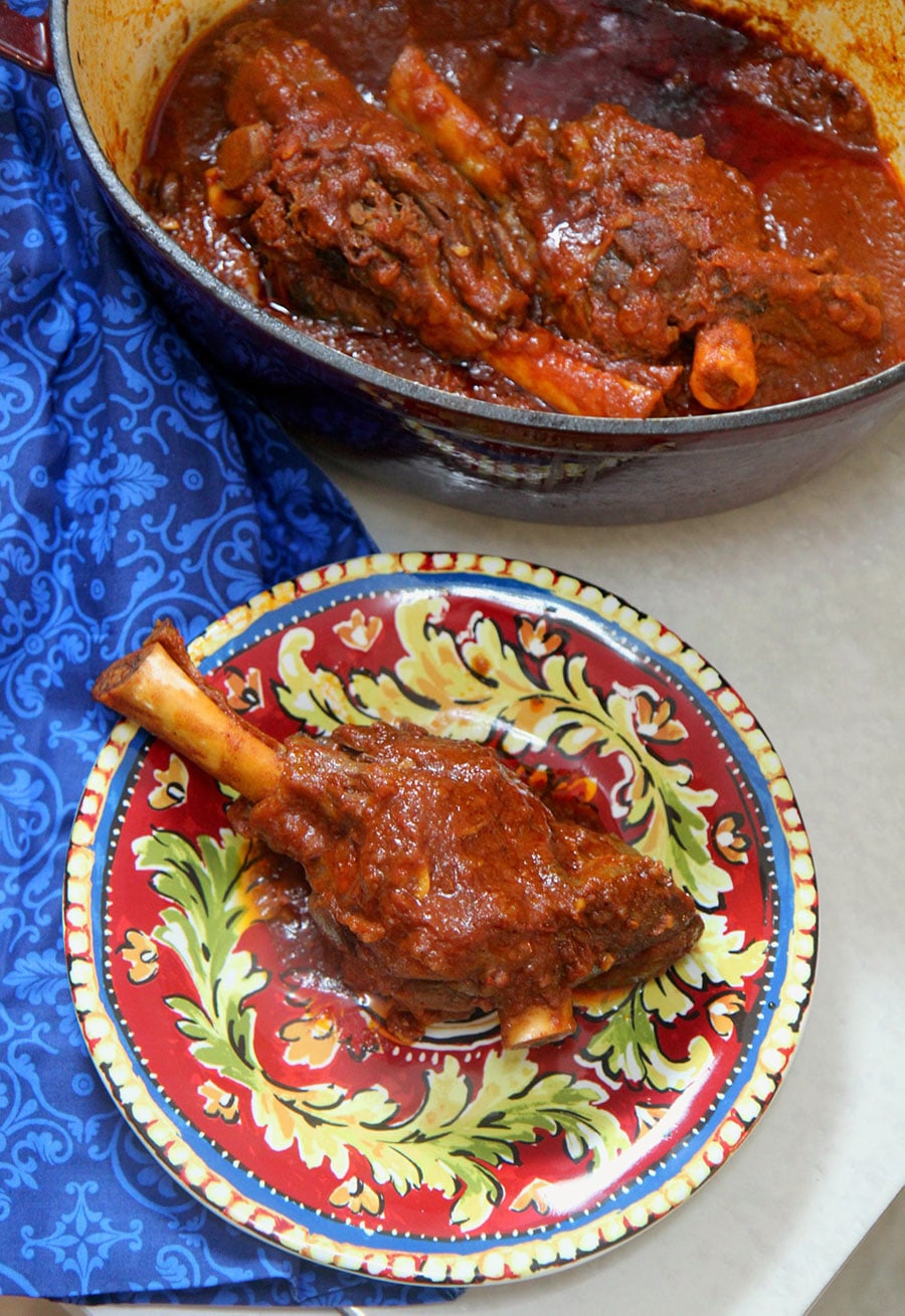 braised lamb shank of a red and yellow plate with a red pot in the background with lamb and a blue towel on the side.