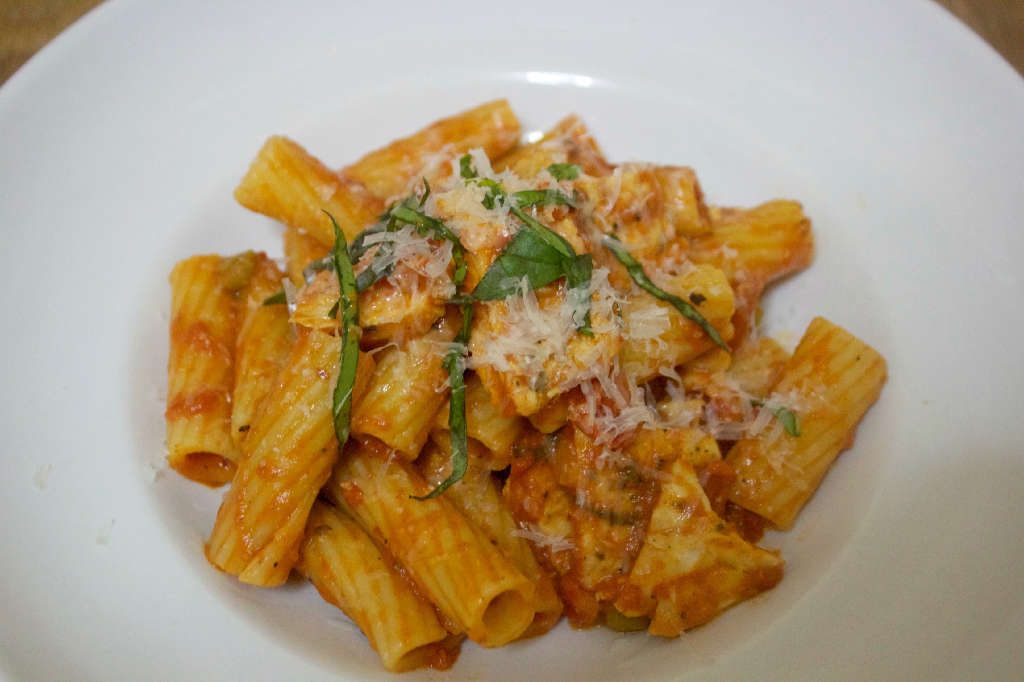 Spicy chicken, rigatoni, parmesan cheese, and fresh basil in a white bowl. 