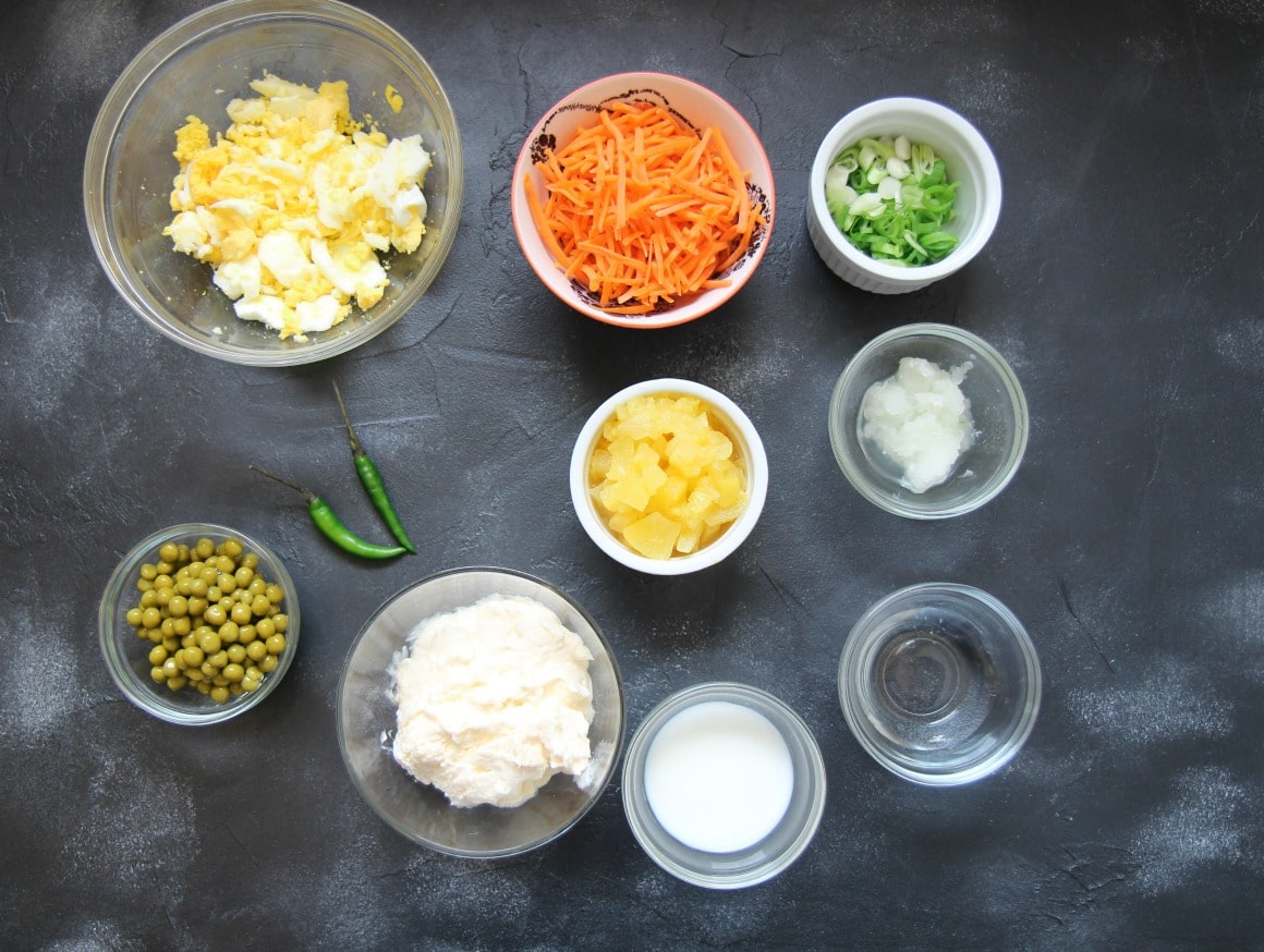 Hawaiian Macaroni Salad Ingredients. Eggs in a bowl, shredded carrots in a bowl, two green chilies, mayo in a bowl, milk, vinegar, scallions, and pineapples in separate bowls. 