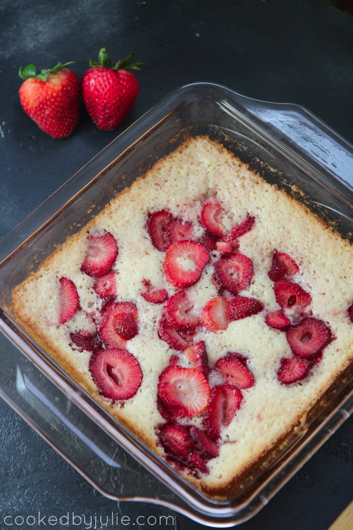 strawberry cobbler in a glass baking dish on top of a black surface. With two strawberries on the side. 
