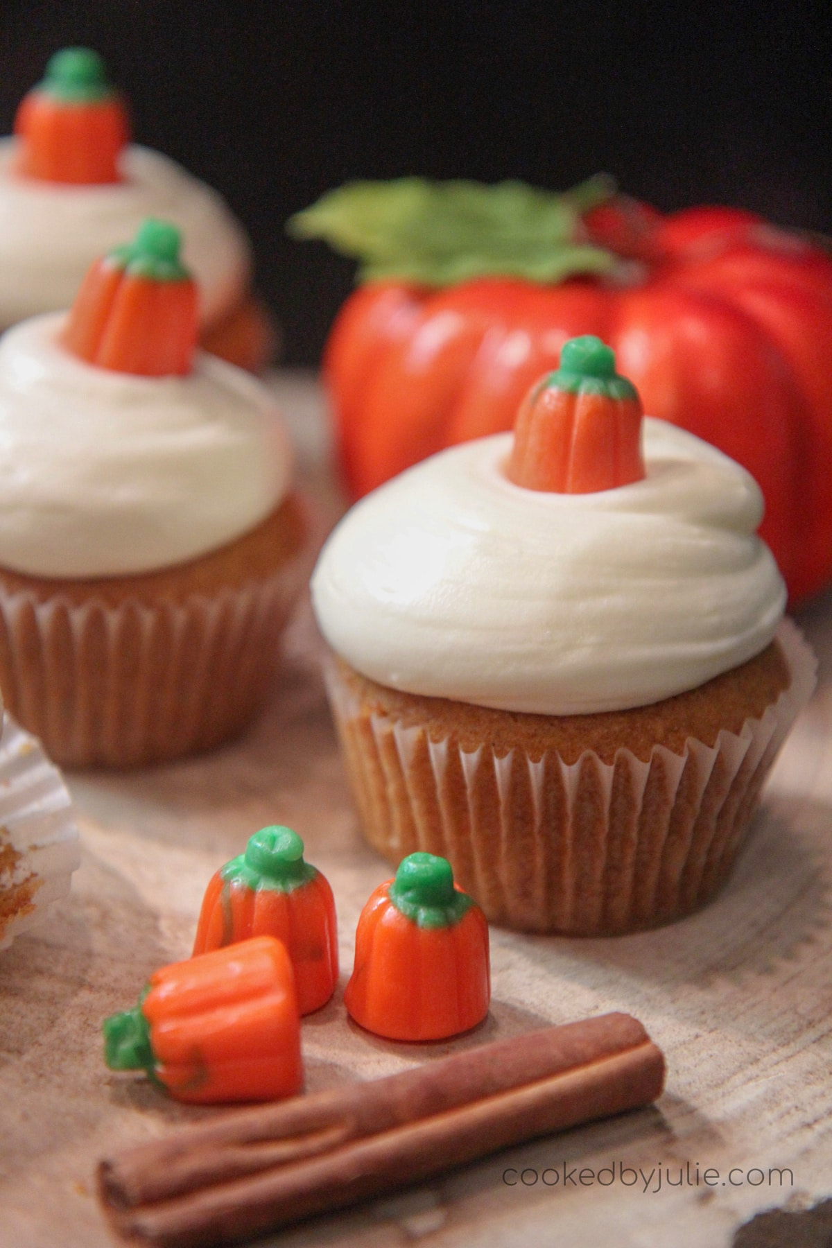 three pumpkin cupcakes with cream cheese frosting with a cinnamon stick, three pumpkin candies, and a fake pumpkin on the side. 